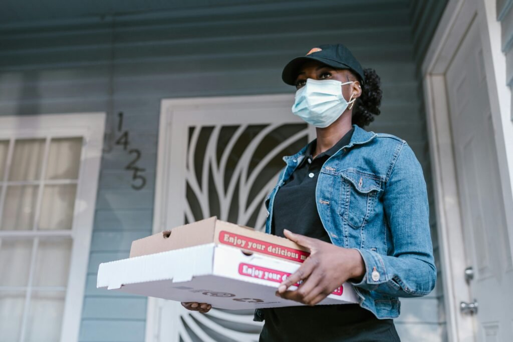 pexels-photo-7363058-7363058 Delivery woman wearing a mask delivering pizza boxes at a front door. Outdoor scene.